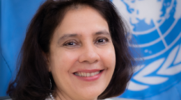 A woman in a white blazer sitting in front of a blue UN flag