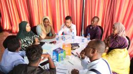 A group of four women and five men gather around a table, in discussion. Papers are on the table along with blocks for the SDG Goals.