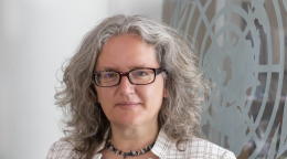 Une femme aux cheveux argentés et aux lunettes foncées se tient devant un mur de verre arborant le logo de l'ONU.