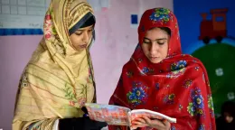 A girl in a yellow headscarf and dress reads a book with another girl in a red headscarf and dress inside a classroom