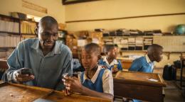 A man in a grey shirt, a teacher, helps a young boy in a school uniform with a mobile device in a classroom.