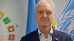 A man in a dark blue suit stands in front of a blue flag