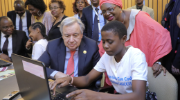 A group of young people along with the Secretary-General Antonio Guterres, peek into a computer as one of them shows them the screen.