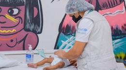 A woman in a grey vest and headcap looks over a baby on a clinic bed.