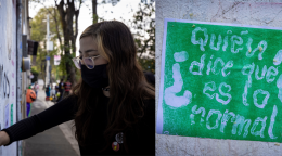 A woman in a black dress and a black mask draws graffiti on a wall.