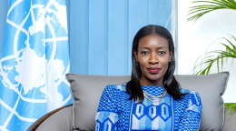 A woman in a blue dress sits on a grey chair against a blue UN flag