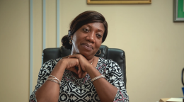 A woman in a black and white blouse sits at a desk with her arms crossed.