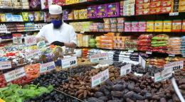 A man in a white shirt and hat, stands behind a cashier in a shop with many commodities and goods.
