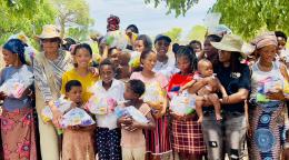 A group of women and children gathered, wearing colourful dresses and hats. In the midst of this group is a woman in a white shirt with glasses and a hat, the UN Resident Coordinator in Namibia