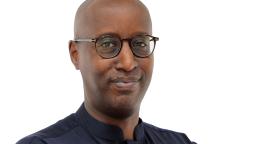 A man in a black shirt and spectacles smiles to the camera as he poses with his arms crossed