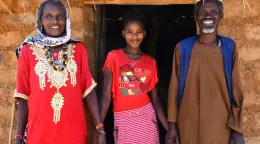 A woman, a young girl and a man stand outside a brick house. They are wearing colourful clothes and smiling.