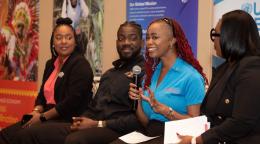 A woman in a blue t-shirt speaks into a microphone while three other people, two women and a man listen intently to her.