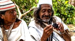 Two indigenous peoples, a man and a woman dressed in traditional clothes sitting side by side, the man in using a mobile phone