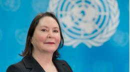 A woman in a black suit and white shirt stands in front of a blue flag with the UN logo on it
