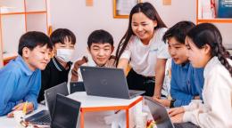 A group of smiling schoolchildren surround an instructor who is pointing at a computer screen.A group of smiling schoolchildren surround an instructor who is pointing at a computer screen.
