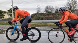Два человека в оранжевых велокуртках едут друг за другом на велосипедах