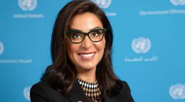 The RC is looking at the camera and smiling. She is wearing a black jacket and glasses and standing in front of a UN blue background with the UN logo imprinted