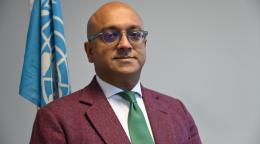 A man in a maroon suit and green tie stands in front of a grey background with a blue UN flag