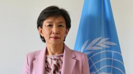 A woman in a pink blazer and shirt stands in front of a blue UN flag