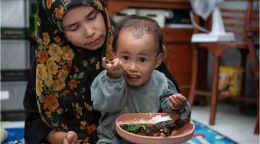 A little boy is eating his food seated in his mother's lap