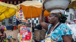 A woman holds a basket of goods on her head, in front of a produce market