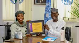 A man in white clothing hands over a commemorative plaque to a woman in a green dress and headdress