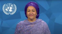 A woman in a purple dress and headdress stands in front of a blue screen with a white UN logo