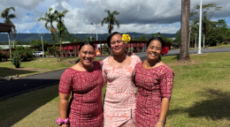 Three people are standing together in colorful dresses in front of a green grassy field with trees