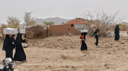 نساء وأطفال في مديرية عبس بمحافظة حجة يحملون صناديق مساعدات غذائية أمام بيوت طينية وأكواخ بسيطة، في مشهد يجسّد معاناة الأسر مع تفاقم الجوع.