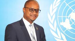 A man in a charcoal grey suit with a grey tie and wearing spectacles stands in front of blue wall with a white UN logo