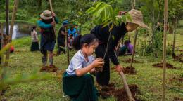 An older woman helps a young child plant a sapling in a lush green mountainous region.