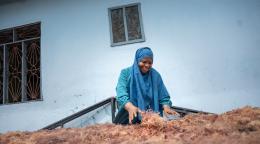 A woman in a blue dress and headscarf sits through piles of brown dried up seaweed.