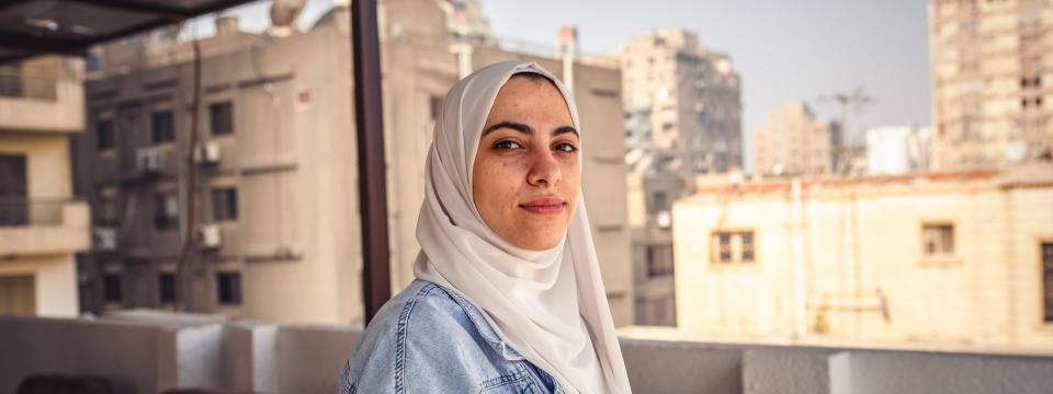 A girl in a white head scarf and denim jacket faces the camera, on a sunny rooftop amid tan-colored high-rise buildings. 