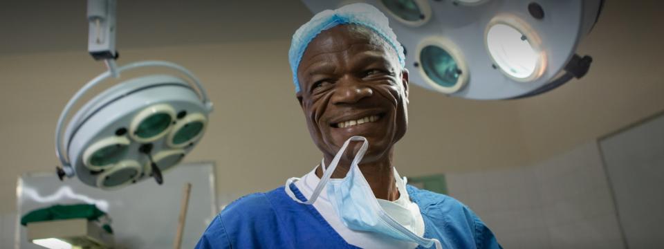 A man surrounded by medical equipment and dressed in a surgical gown smiles and holds a mask by his teeth.