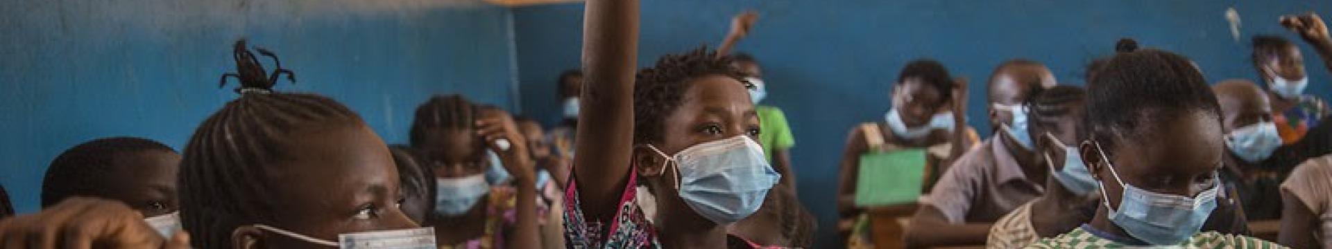 In a classroom of students wearing face masks, one student proudly raises their hands.