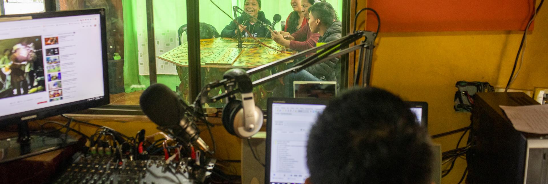 Four young people speak into a microphone in a green room in front of a person with 
