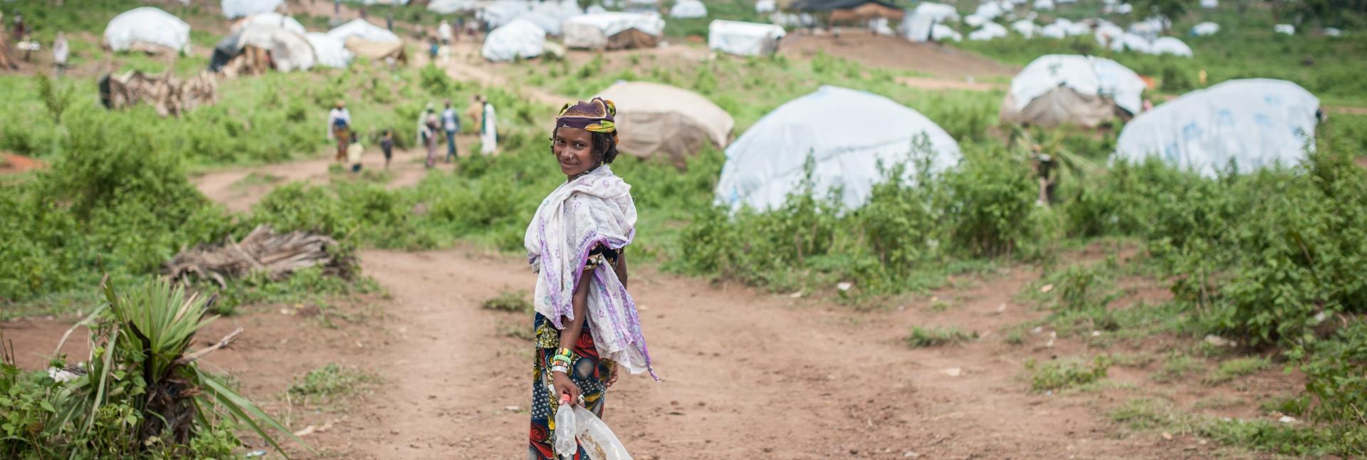 A woman turns back and smiles at the camera as she walks towards a green patch, with tents dotting the horizon.