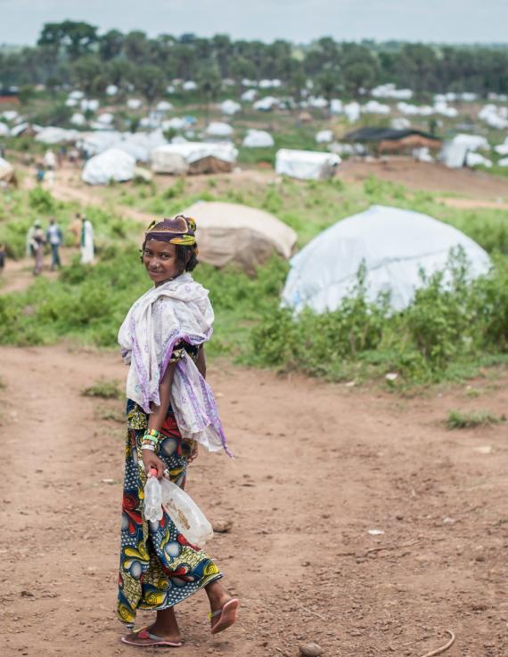 A woman turns back and smiles at the camera as she walks towards a green patch, with tents dotting the horizon.