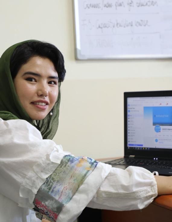 A young Afghan woman smiling at the camera, with a laptop on the desk in the background. 