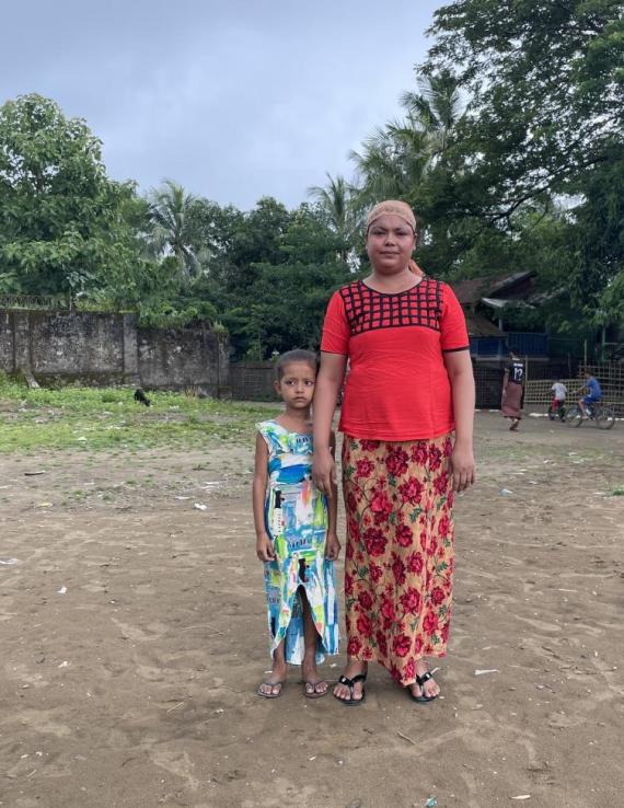 A woman stands next to a young girl, on a dry patch of land with some grass, trees, and people on motorbikes behind them.