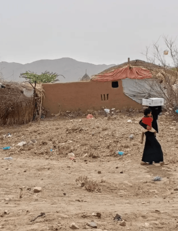 نساء وأطفال في مديرية عبس بمحافظة حجة يحملون صناديق مساعدات غذائية أمام بيوت طينية وأكواخ بسيطة، في مشهد يجسّد معاناة الأسر مع تفاقم الجوع.