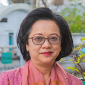 A woman in a pink colored silky top and glasses smiles on a sunny day outside and faces the camera.