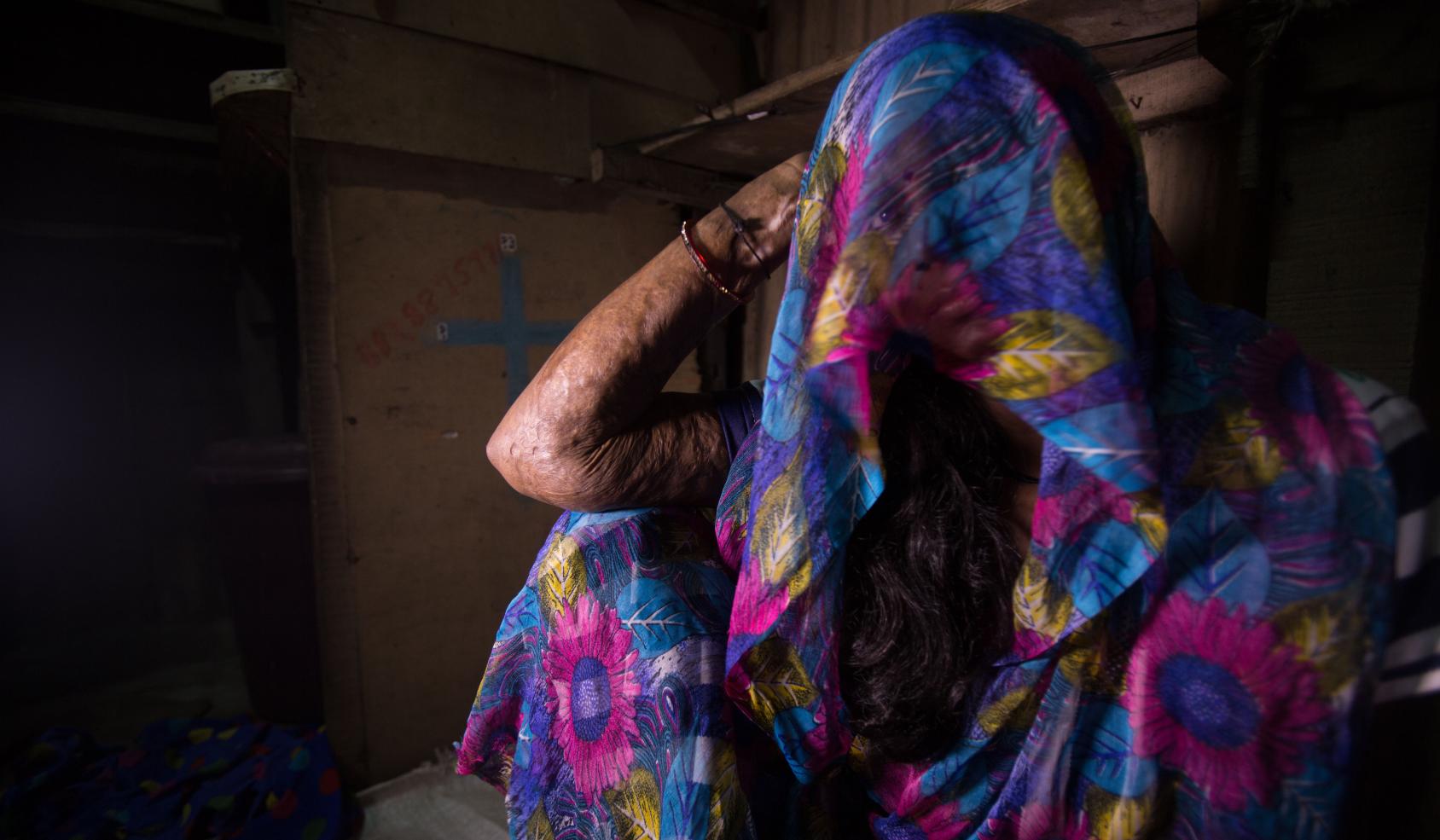 A woman sits on the floor and looks at the camera with the fabric from her dress over her head covering her face.