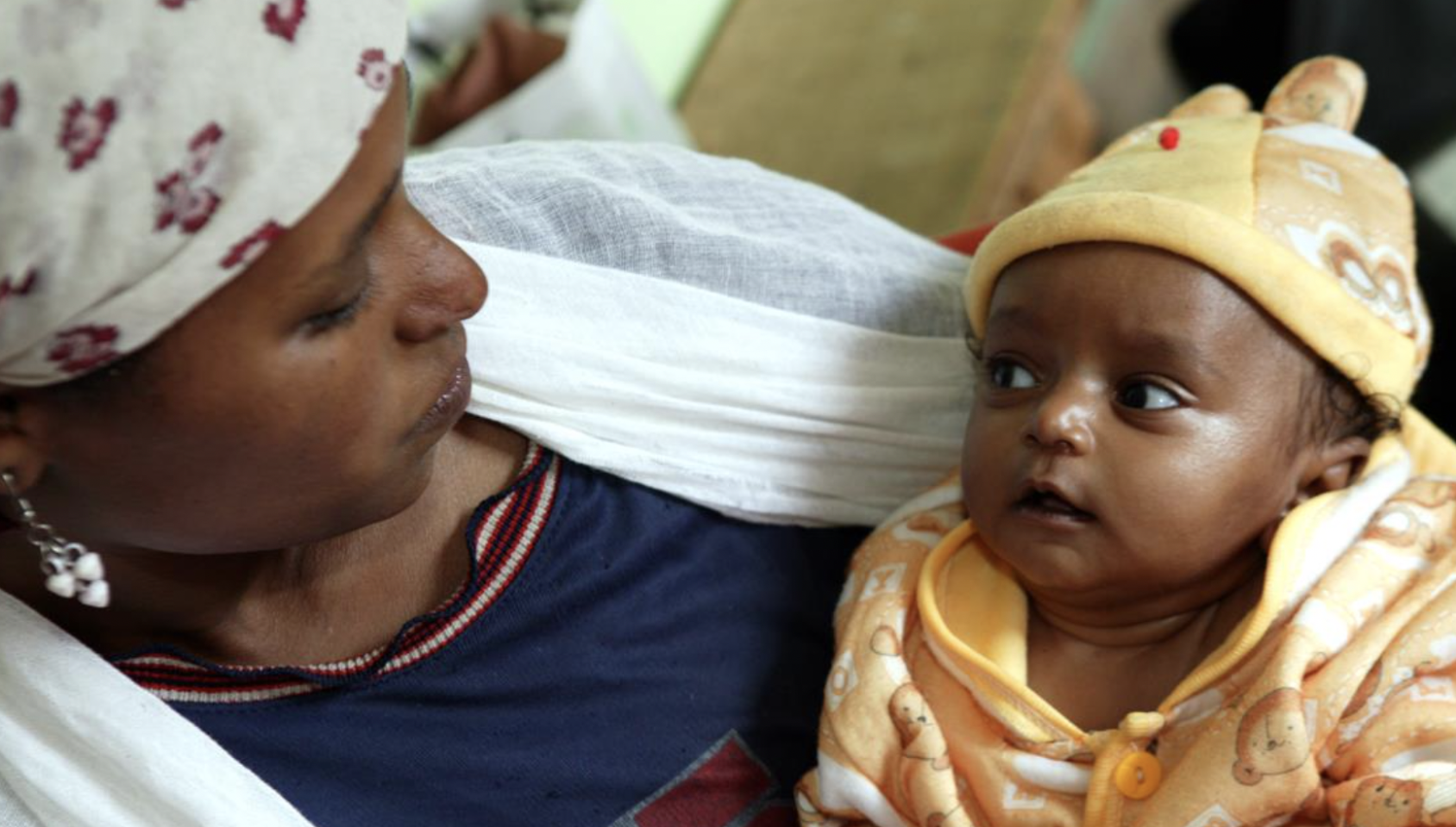 Une femme tient son bébé dans les bras en le regardant. L'enfant est vêtu d'un pull jaune et d'un bonnet jaune et regarde fixement sa mère.