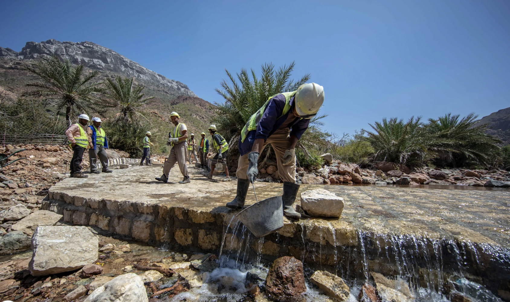 Des ouvriers travaillent à la remise en état d'une route au milieu d'un paysage fait de montagnes et de palmiers. Au premier plan de l'image, un ouvrier se penche en avant pour déverser le contenu d'un seau.