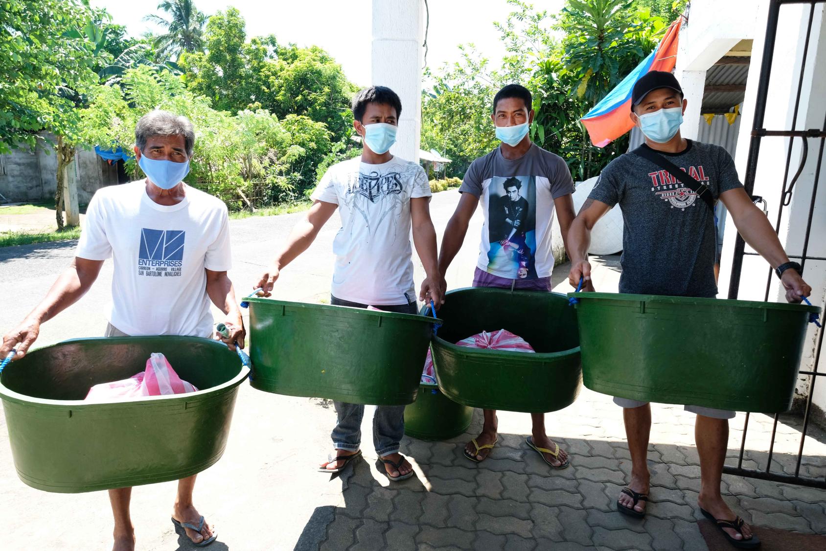 Quatre hommes vêtus de t-shirts et portant des masques de protection respiratoire se tiennent debout, côte à côte, sur la pallier d'un bâtiment, chacun tenant entre les mains une grande bassine verte contenant du matériel de pêche.