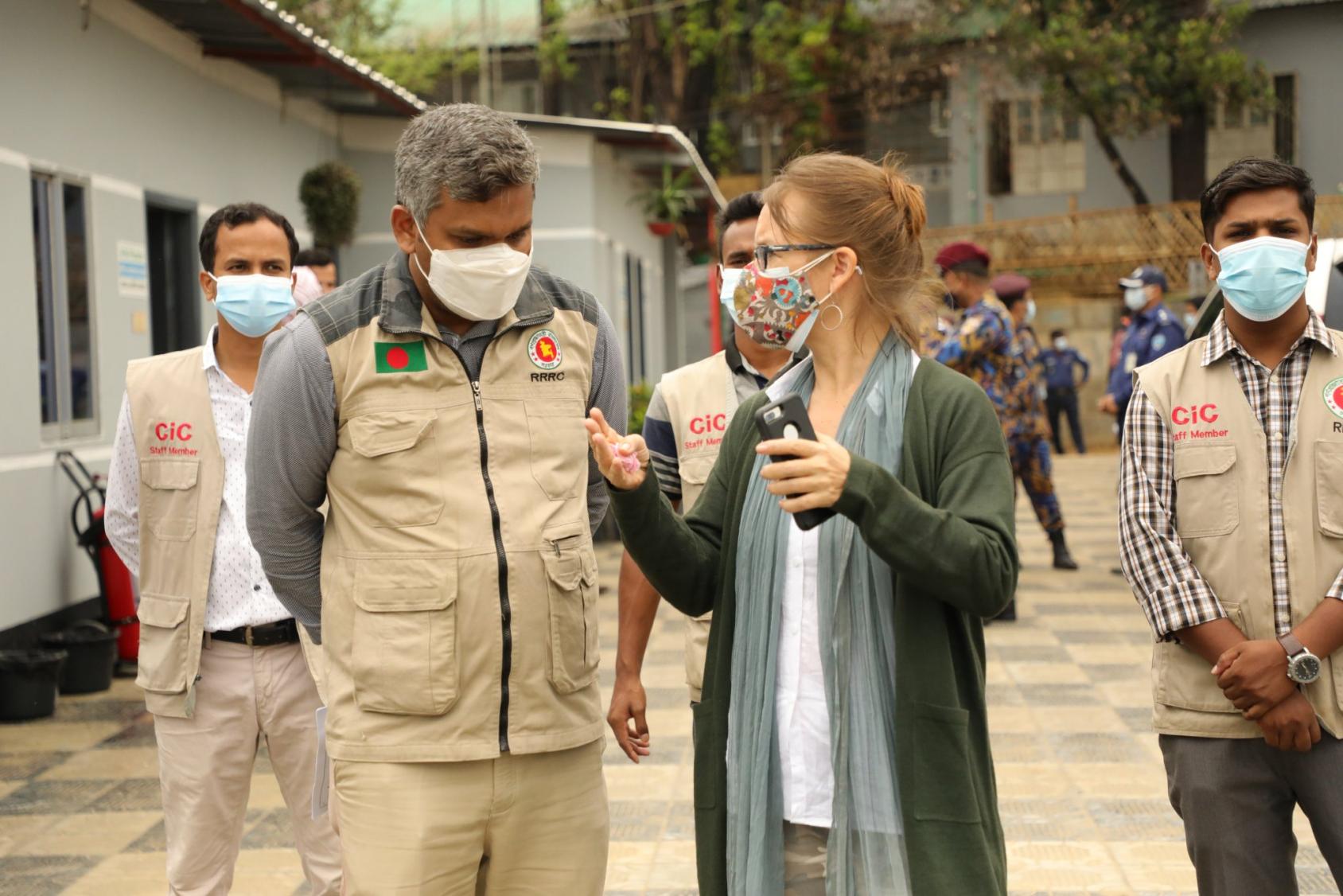 Une femme tenant un téléphone portable à la main gauche s'entretient avec un homme aux cheveux grisonnants qui a les bras croisés derrière le dos. Trois hommes se tiennent debout derrière eux, le regard fixe. Toutes les personnes présentes sur la photo portent un masque de protection respiratoire. 