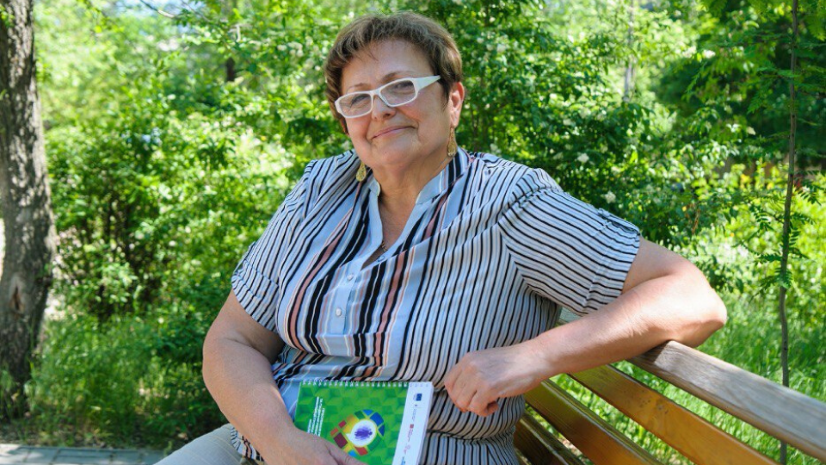 A woman, surrounded by nature, sits at a bench with a book in her hand.