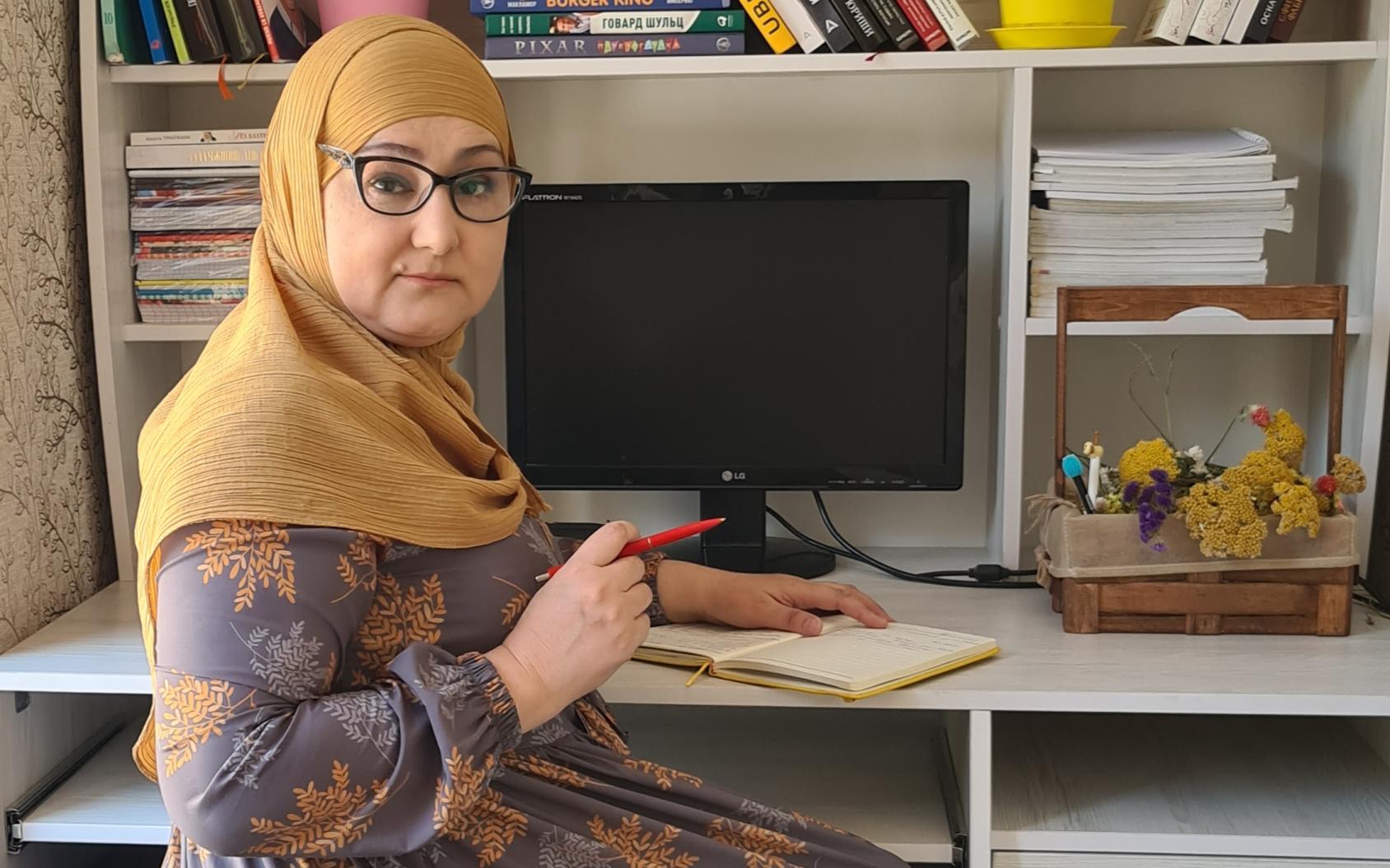 Une femme portant des lunettes et un foulard sur la tête et autour des épaules est assise à un bureau, chez elle, et tourne le regarde vers l'objectif. Sur son bureau son posés un cahier, dans lequel elle s'apprête à prendre des notes, ainsi qu'écran d'ordinateur et des étagères garnies de livres.