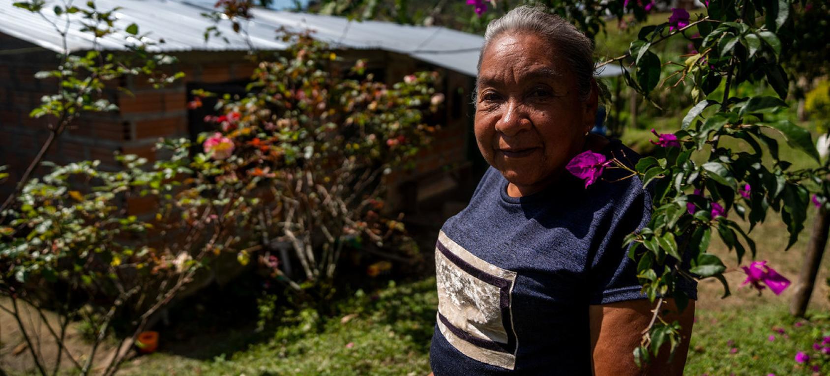 Una mujer mayor mira directamente a la cámara, mientras se encuentra parada cerca de vegetación en flor.